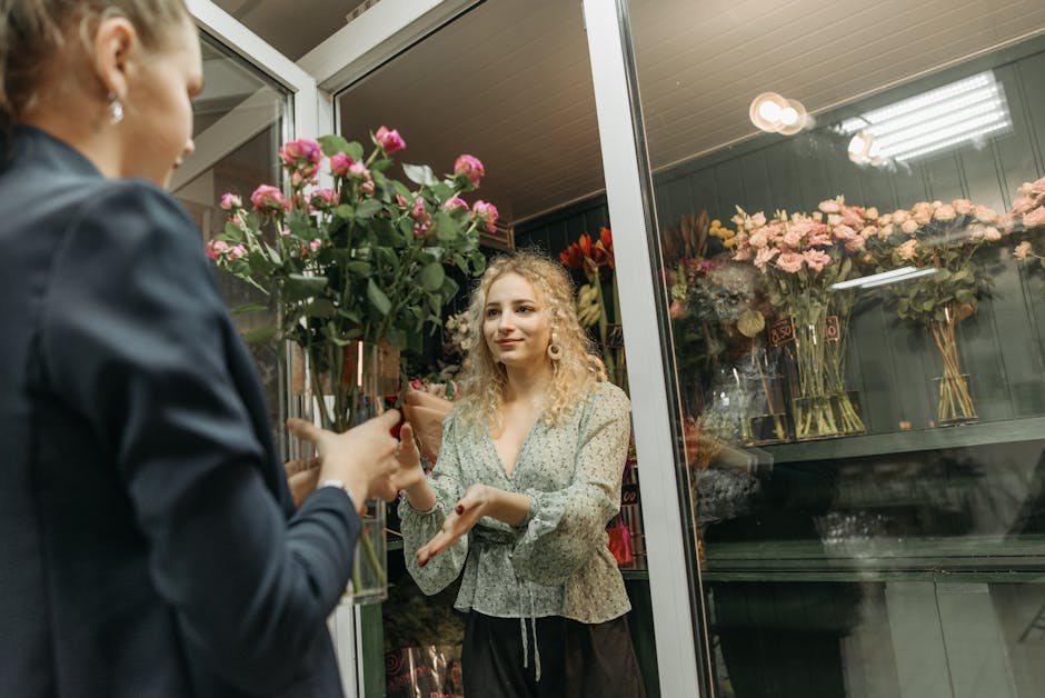 A florist with curly blonde hair, wearing a light-colored, patterned blouse, hands a bouquet of pink roses to a customer through a glass shop door. The bouquet is arranged with numerous pink roses, gr