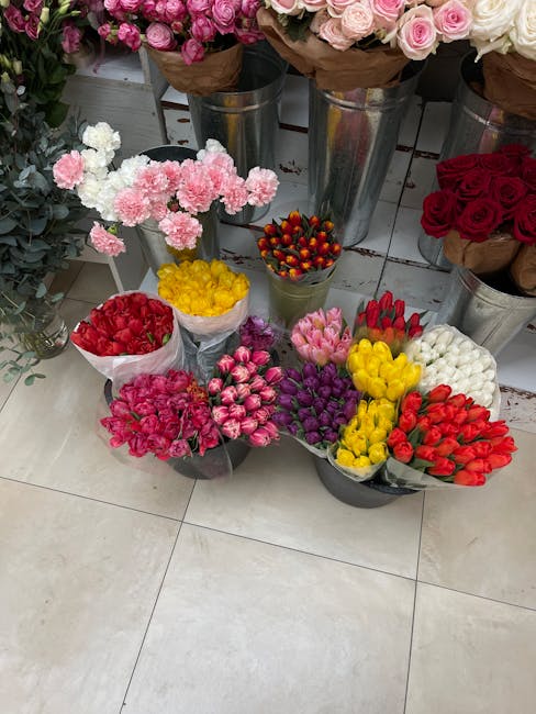 A display of various fresh flower bouquets and individual flower bunches arranged on a tiled floor in a floral shop. The flowers include pink carnations, red roses, white roses, yellow tulips, purple