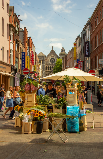 A vibrant outdoor flower market scene on a busy city street, showcasing a colorful display of various fresh flowers arranged in wooden crates and buckets. The flowers include sunflowers, roses, lilies