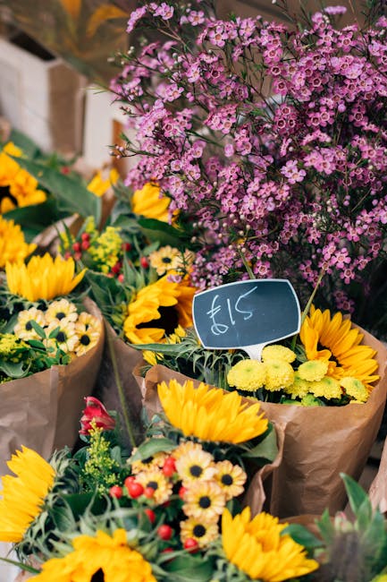 A vibrant floral display featuring a variety of fresh flowers arranged in brown paper wrapping. Prominent yellow sunflowers with dark centers, smaller yellow daisies, and red and white blossoms are vi