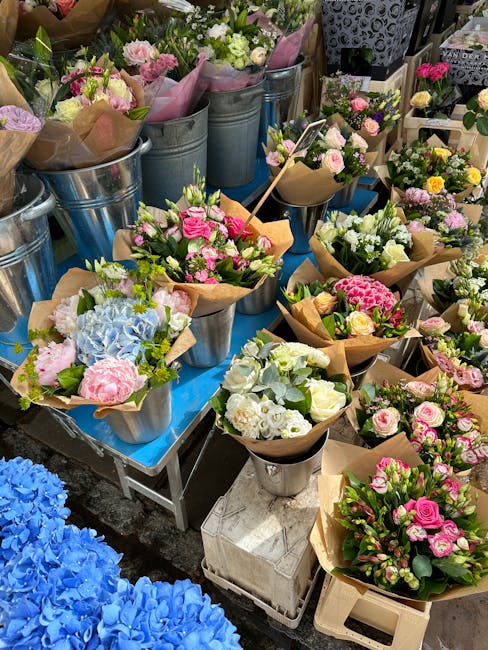An outdoor display of fresh flower bouquets arranged in metallic buckets and wrapped in kraft paper, featuring pastel-colored roses, hydrangeas, and carnations in shades of pink, white, and green, wit
