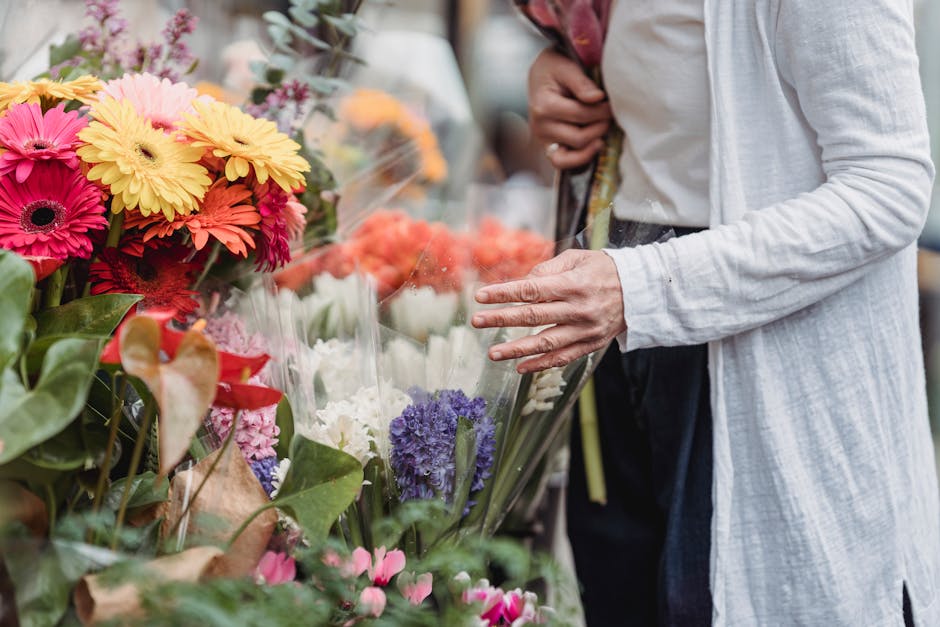 A person dressed in a light gray long-sleeve cardigan and dark trousers, selecting a bouquet of fresh flowers at a flower market. The arrangement includes brightly colored gerbera daisies in shades of
