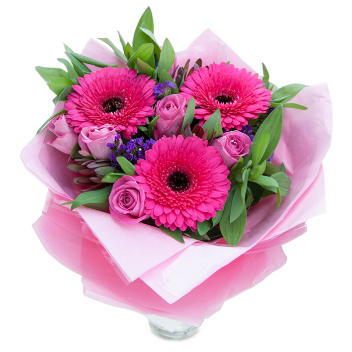 The charm of this pink bouquet is quiet and composed: three fuchsia Gerbera daisies open like joyful faces, their intense colour softened by the deep, almost black centres that lend a touch of drama. Close to them, roses in a dusty lavender-pink keep their petals gently wrapped, an intimate counterpoint to the Gerberas' exuberance. Interspersed among the main blooms are small clusters of purple statice, their papery texture and cool hue introducing a thoughtful punctuation to the overall rhythm. Rich green foliage-leaves that resemble salal or eucalyptus-creates a garden-like base, its matte surface and subtle scent of green leaves giving the bouquet a sense of grounded freshness. The stylistic wrapping is part of the story: an inner layer of soft pale-pink tissue that cradles the blooms, and an outer sheath of translucent, slightly crinkled fuchsia-pink cellophane that catches light and ties the colours together. At the lower edge the top of a clear glass vase peeks through, suggesting immediacy-this is a bouquet arranged to be seen and enjoyed at once. Under a gentle, late-afternoon glow the petals hold soft highlights and shadows, and the faint perfume of roses blends with a green, leafy aroma. I imagine this piece brightening a mantel in a cottage near the River Lea or adding a warm note to a table by Lowewood Museum in Hoddesdon. It's a composed, tender choice for anniversaries, Mother's Day, birthdays, or simply to convey affection with understated elegance.